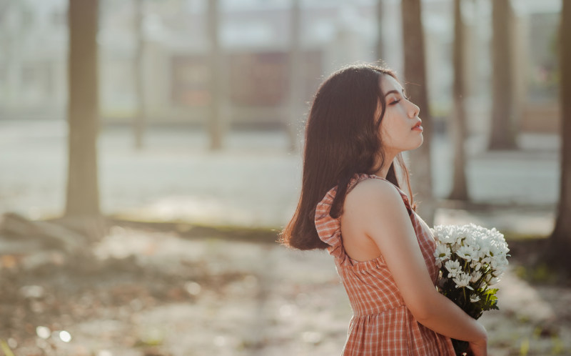 一个女人，穿着棕色和白色的勺子领无袖连衣裙，手里捧着一束白色的花瓣花|侧视图,公园,头发,女人,女孩,户外,日光,时尚,服装,树干,模糊的背景,花束-海量高质量免版权图片素材-设计师素材-摄影图片-mitapix-美塔图像